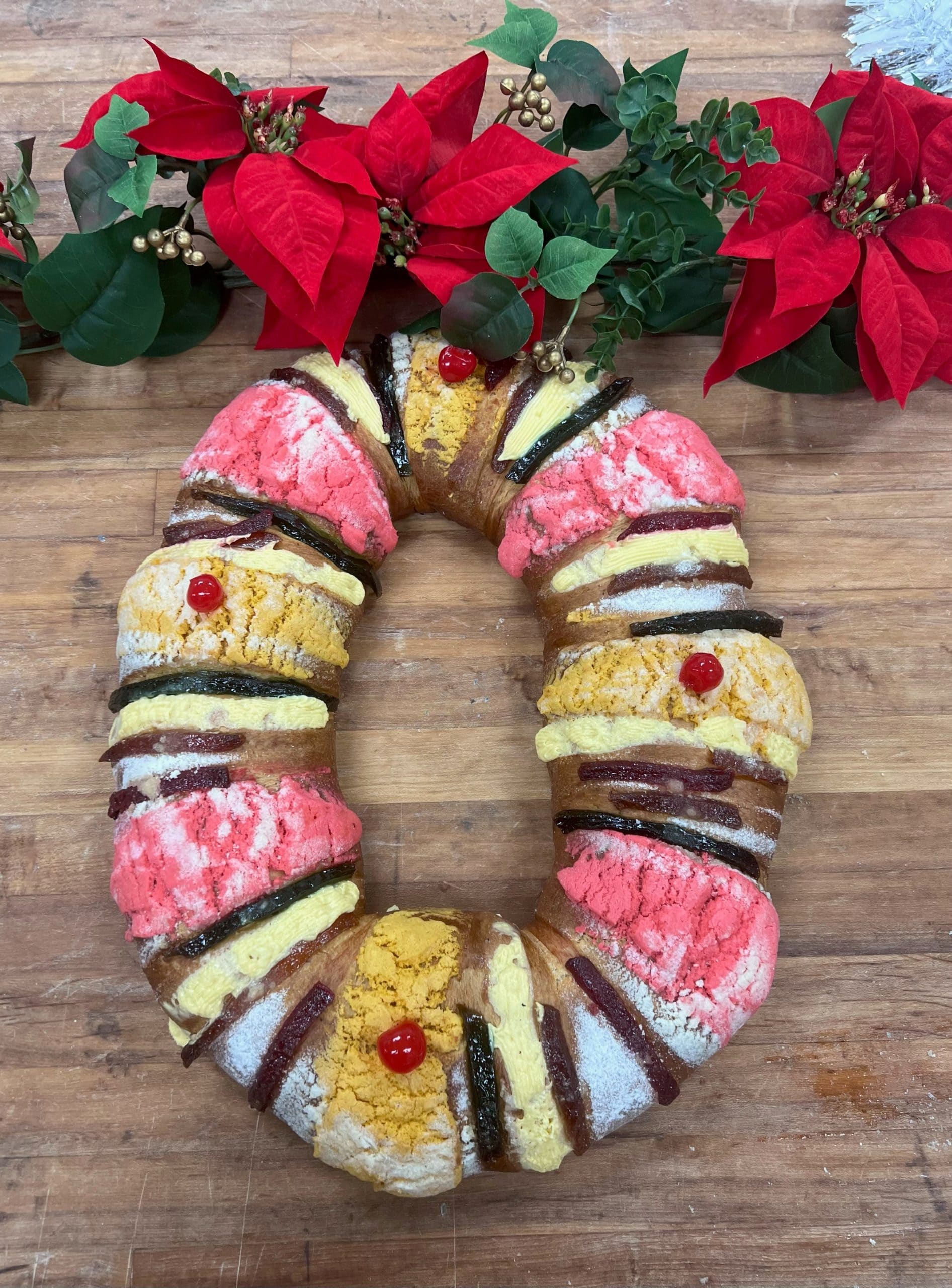 A brightly decorated Rosca de Reyes sits on a wooden surface, surrounded by festive poinsettias.