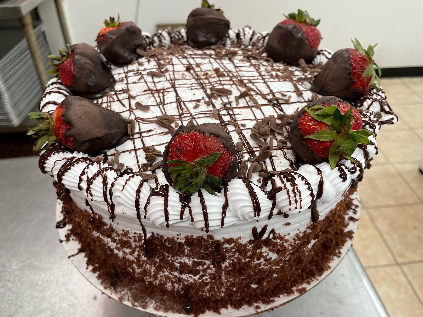A chocolate cake is elaborately decorated with white frosting, chocolate drizzle, chocolate crumbs, and several chocolate-dipped strawberries.