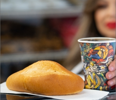 A person holds a colorful coffee cup next to a golden-brown bread roll.