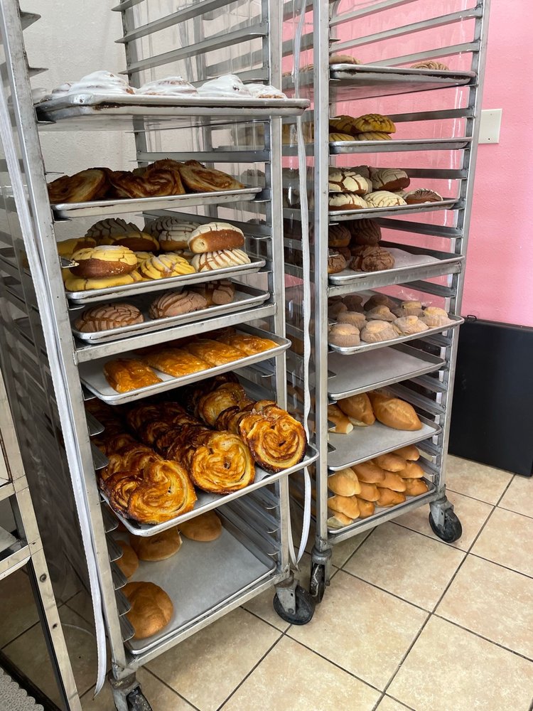 Two multi-tiered metal racks are laden with a wide variety of baked goods, including pastries and breads, in a bright pink-walled bakery.